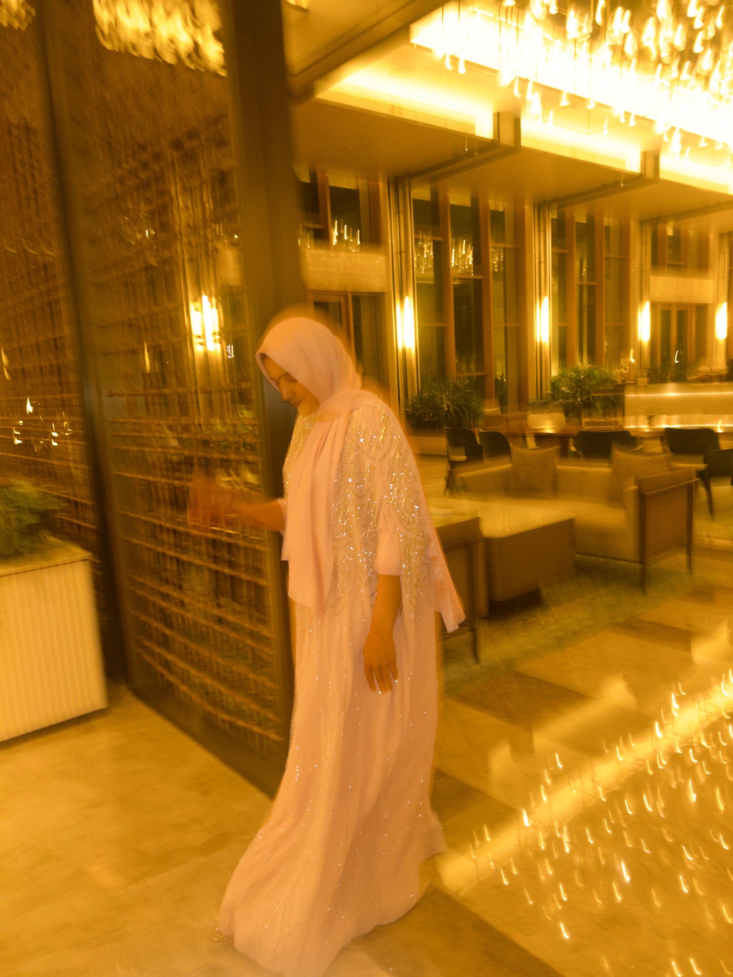 Woman in a white dress standing in a luxurious interior setting with chandeliers and seating.