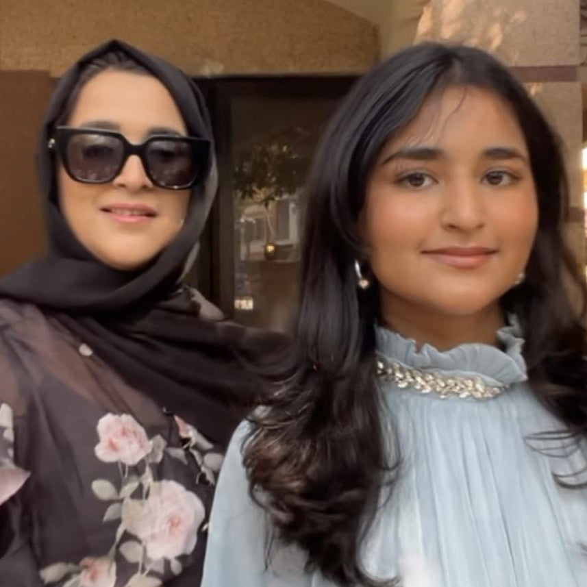 Two women standing together outdoors, one wearing a floral dress and sunglasses, the other in a light blue dress.