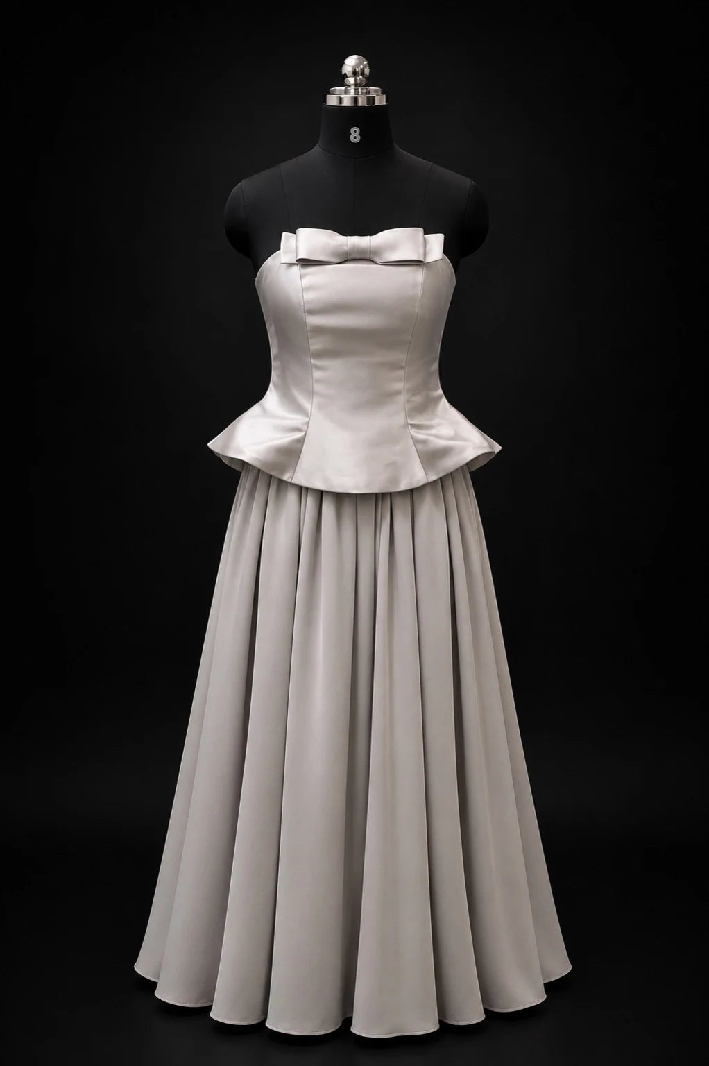 A strapless formal gown displayed on a black mannequin against a black studio background. The dress has a structured corset-style bodice with a bow at the bust, a peplum waist, and a full flowing skirt.